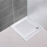 White plastic, square shower mat in a slatted design in situ on a plain white, square shower tray in the corner of a bathroom with a white floor and one grey tiled wall and one plain white wall