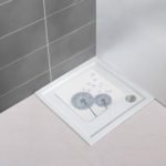 a white, square shower mat with grey silhouettes of two dandelion clocks with a few seeds being scattered in situ on a plain white, square shower tray in the corner of a bathroom with a white floor and one grey tiled wall and one plain white wall