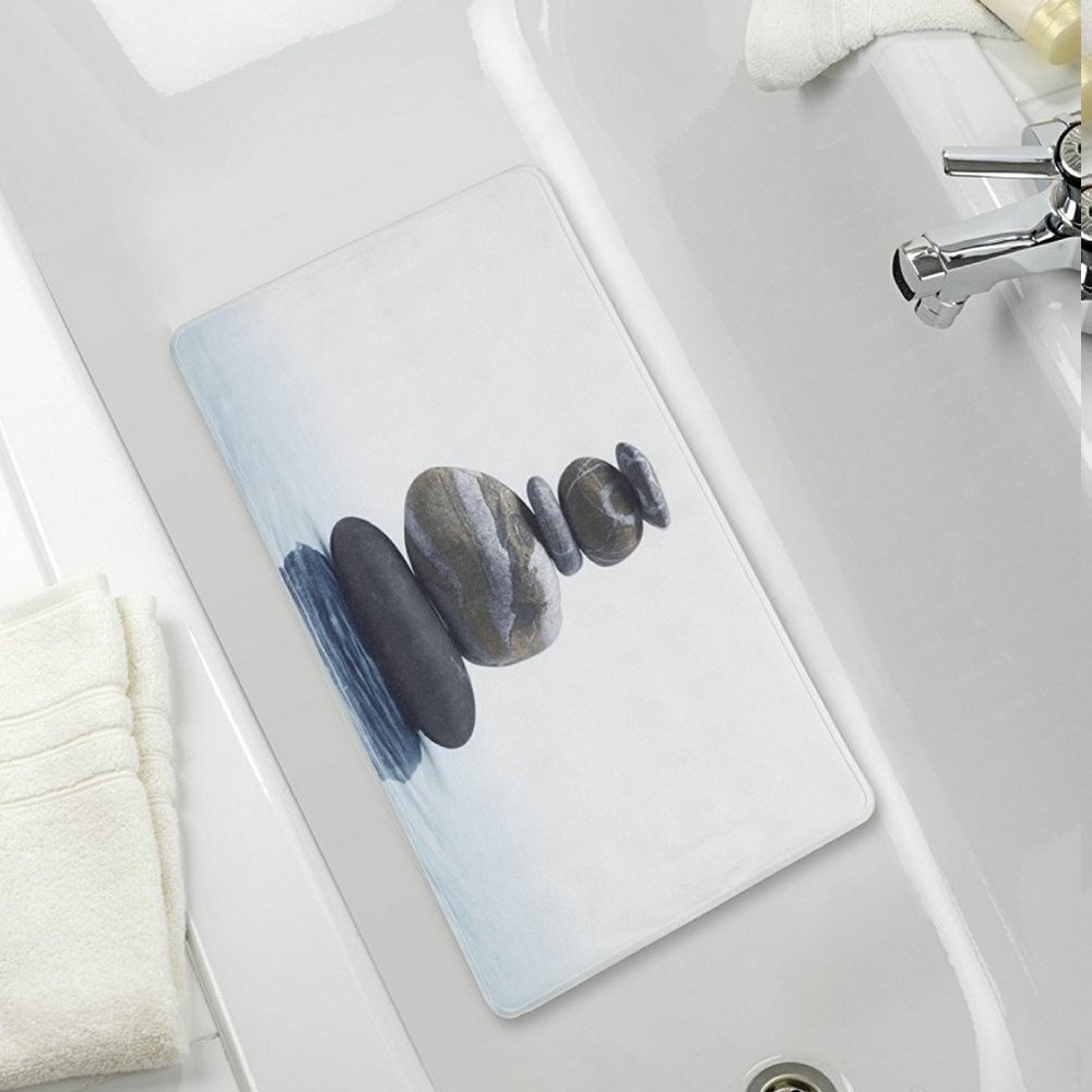 White, rectangular bath mat with a slight blue-grey gradient on the bottom with ripples, indicating water. In the centre is a neat stack of five grey pebbles balanced on top of each other, the two on the bottom are somewhat wider than the top three. It is centred in a white bathtub