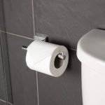 chrome toilet roll holder held onto wall by square bracket on one end, on the other a small protusion stick ot to prevent the roll sliding off. it is on a gray tiled surface, next to a white cistern and is holding a toilet roll