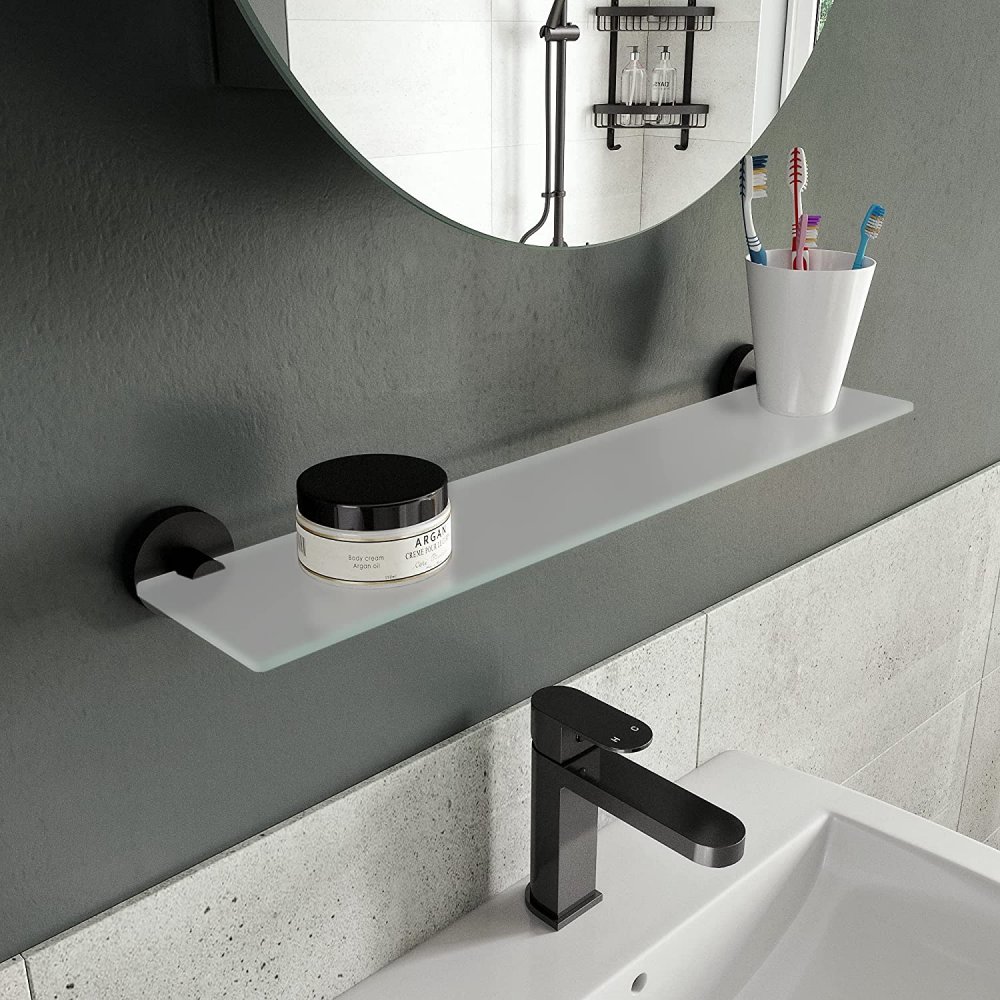 glsss shelf with matt back wall brackets on each endit is on a grey wall under a round mirror and above a white sink with a single basin ixer. on the shelf is a small jar with a black lid and a white tumbler containing tooth brushes