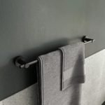 matt black towel rail with round wall brackets on each end, it is on a grey wall and is holding a grey towel and another smaller grey towel