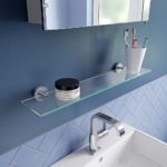 rctangular glass shelf with a round chrome wall bracket on either end. It is on a blue wall under amirrored cabinet and above a white sink with a chrome mixer tap. On the shelf is a wjite jar with a back lid and a white tumbler with toothbrushes in