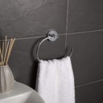 chrome towel ring with round back plate. the towel ring is in an incomplete oval shape.. it is on a grey tiled wall holding a white towel by a white sink with a reed diffuser placed on it