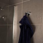 chrome double robe hook, it is on a dark grey tiled wall next to a glass shoer encloser and holding a dark blue towel
