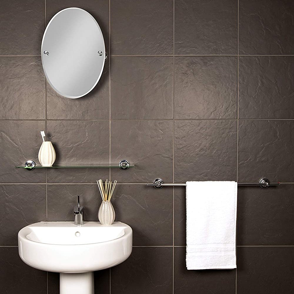 on the left hand side of the image is a round mirror with chrome wall brackets. it is mounted on a dark grey tiled wall above a glass shelf with white oval tumbler containing a single toothbrush which, in turn is above a white sink featuring an oval reed diffuser. on the right had side of the image is a straight towel rail holding a white towel.