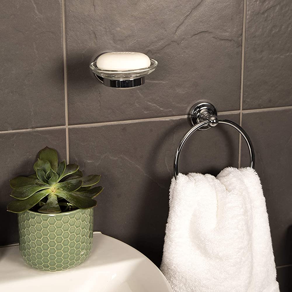 chrome towel ring with round back plate. the towel ring is in a circle shape it is mounted on a dark grey tiled wall and holding a white towel, it is to the right of a white sink with a green succulent in a pale green vase on the back. Above and to the middle of the two is a glass soap dish on a chrome mount holding a white bar of soap